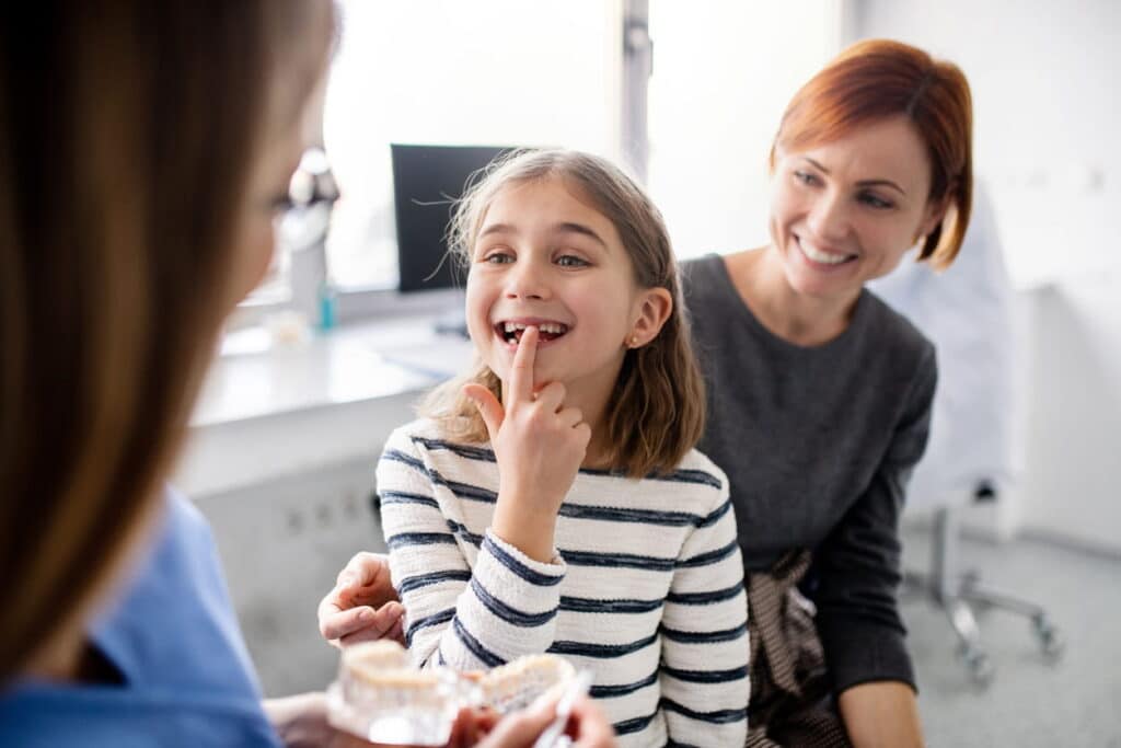 A daughter smiling, showing her missing tooth to the family dentist. Behind her is her mother, also smiling.