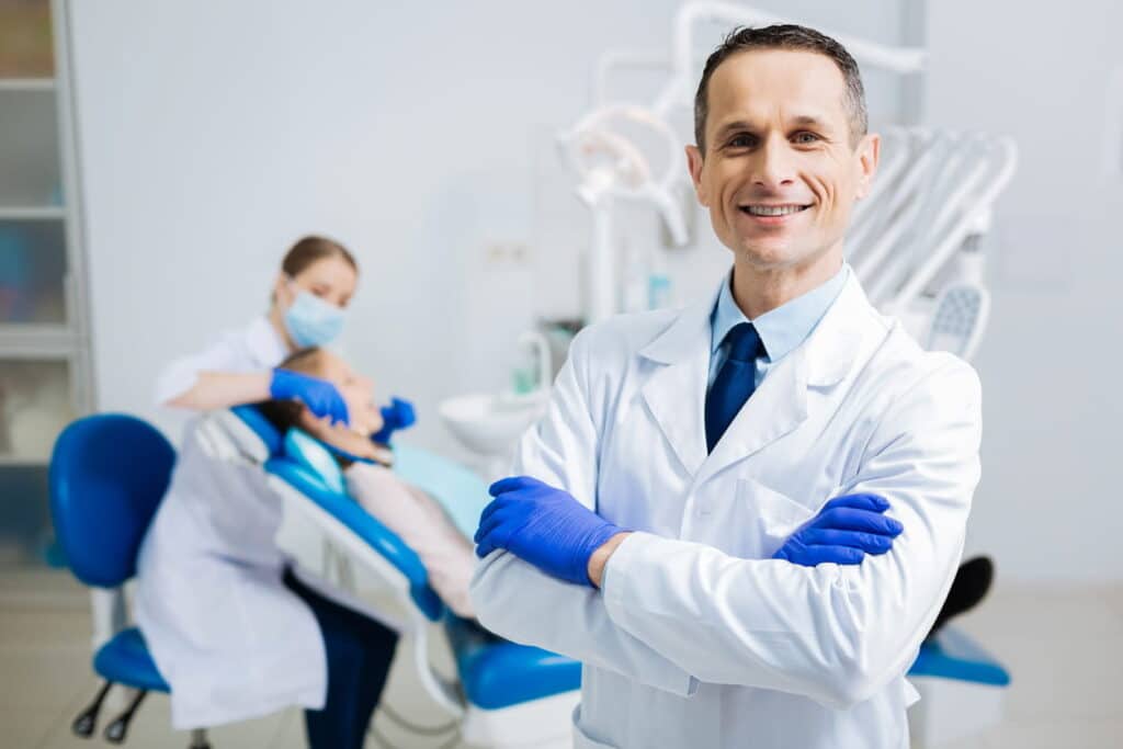 A male family dentist in Las Vegas smiling. In the background a dental hygienist is working on a patient.