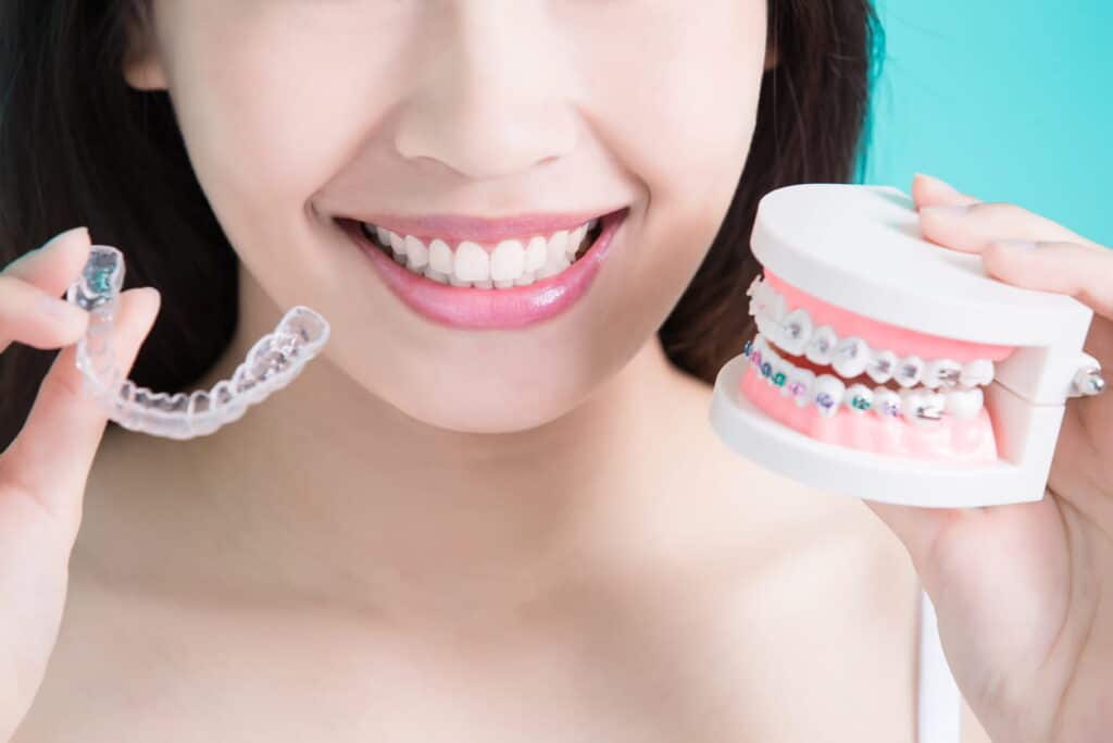 Focus is on a woman smiling as she holds clear aligners in one hand and a model of a mouth with traditional braces in the other.