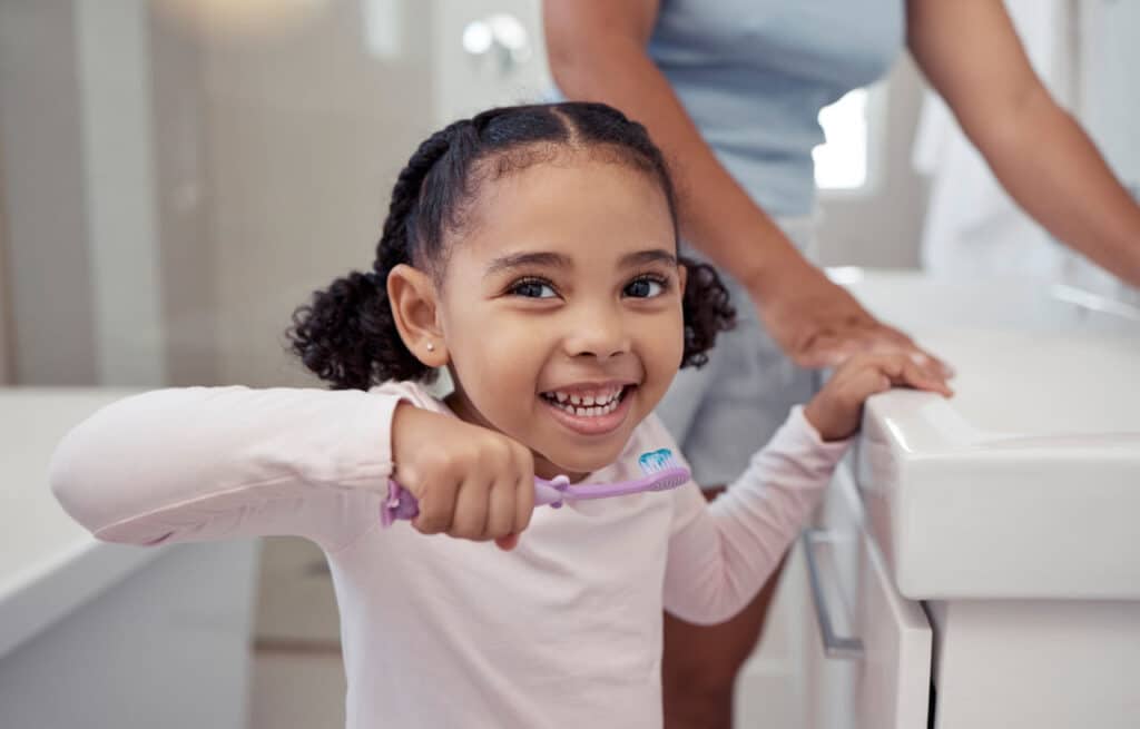 A child smiling and brushing her teeth before seeing a children's dentist.
