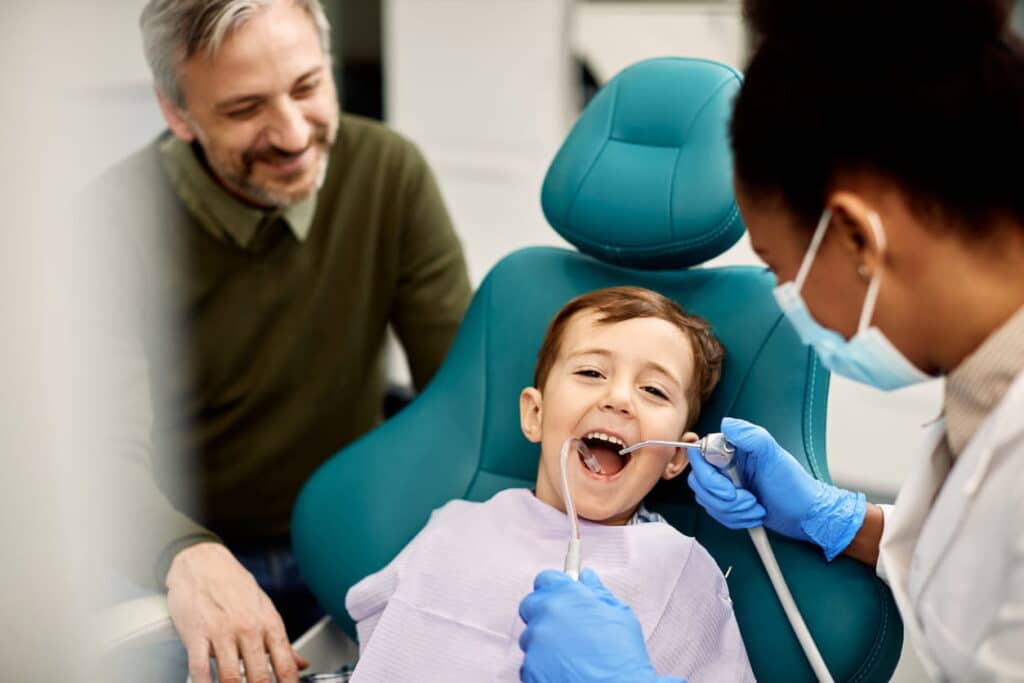 A children's dentist working on their patient who's smiling in the dental chair. His dad is next to them also smiling.