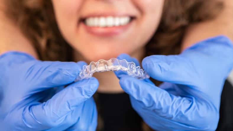 A dentist is holding a clear aligner in front of a patient's mouth as she smiles.