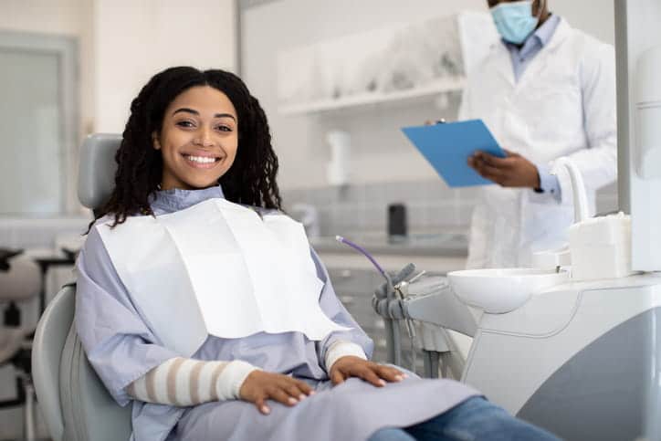 A woman smiling at the dentist's office with the dentist in the background reviewing her chart.