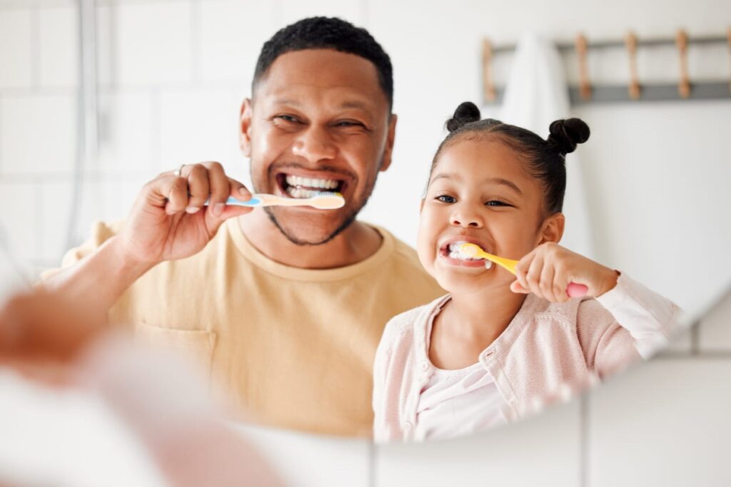 A father and daughter brushing their teeth while smiling in mirror, making fun dental care.