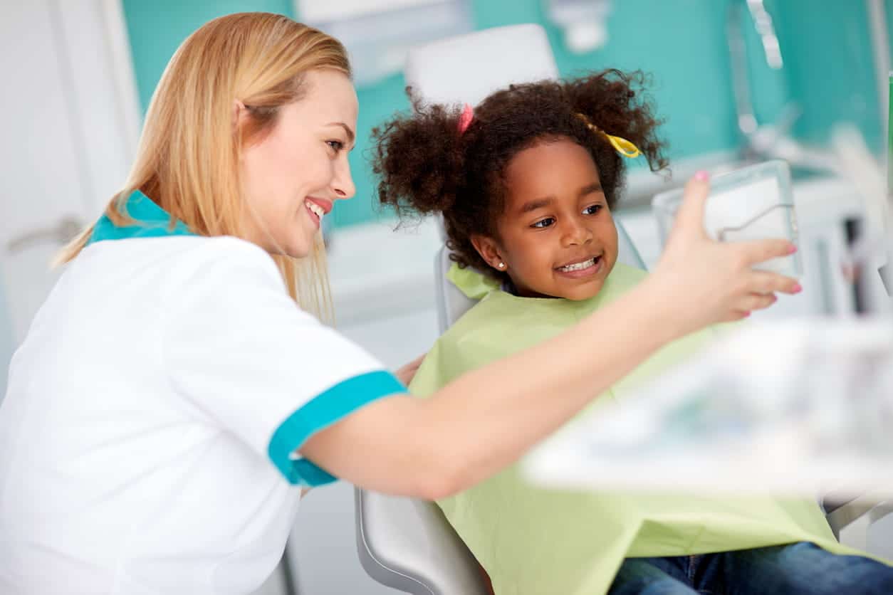 A little girl smiling into a mirror held up by her dentist in Henderson, NV.
