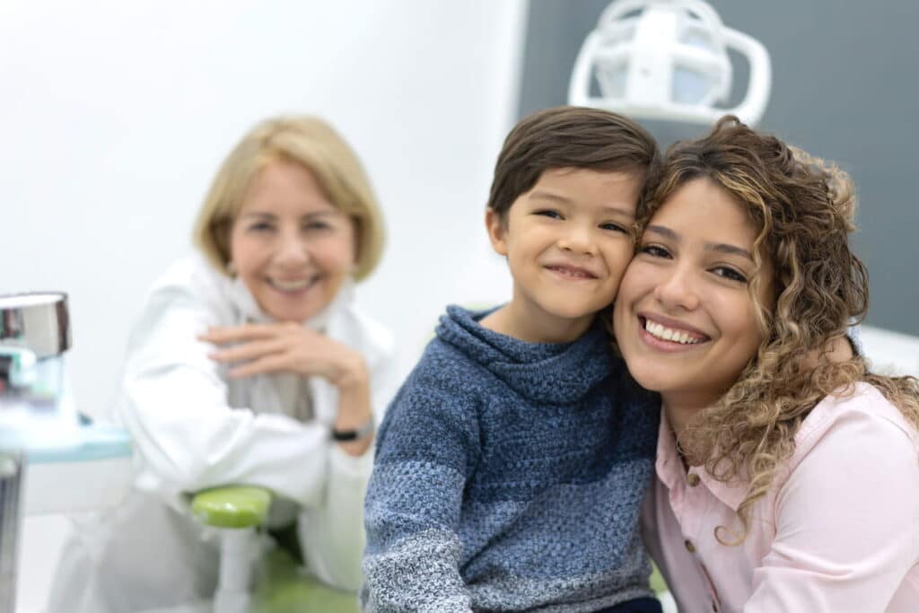 A mother and son smiling while at the dentist's office. The dentist is blurred out in the background, smiling as well.