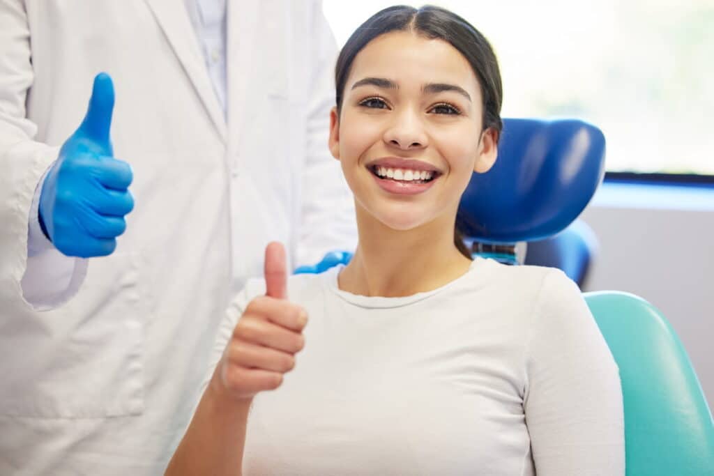 A woman at the dentist's office giving a thumbs up. The dentist, mostly out of frame, is also giving a thumbs up.