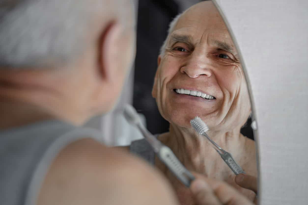 An elderly man smiling into the mirror with dentures. He's holding a toothbrush in his hand.