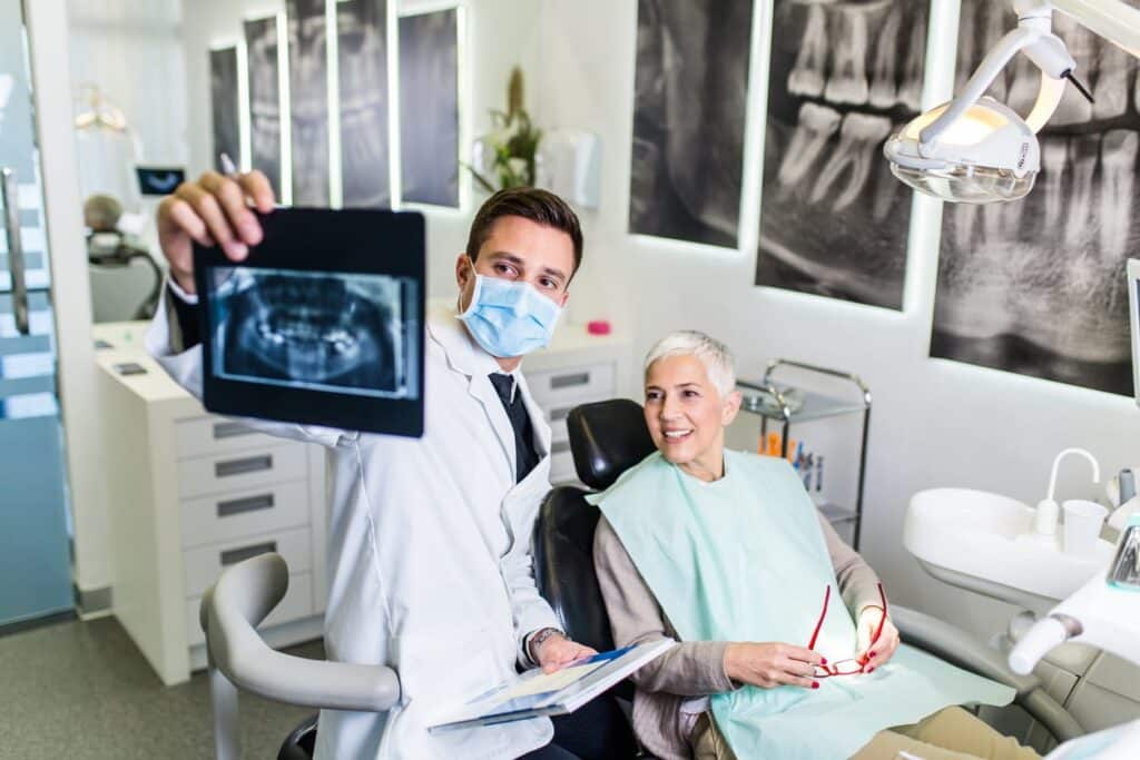 A geriatric dentist in Sparks, NV is showing his elderly patient an X-ray of her mouth. She's sitting in the chair smiling while holding her glasses.