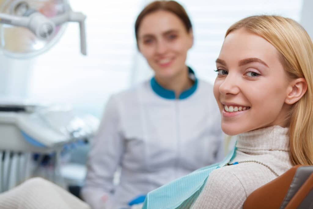 A woman in a dental chair smiling after being seen by a Las Vegas dentist for a dental emergency. The dentist is blurred out in the background. 