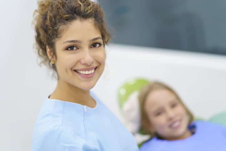 A dentist smiling for the camera as her young patient smiles in the background, blurred out.
