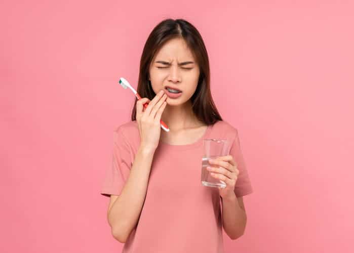 A teenager with braces is holding her face in pain while holding a toothbrush and a glass of water. 