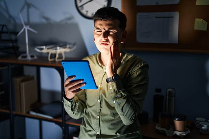 An individual holding their face in pain from a toothache while on their tablet.