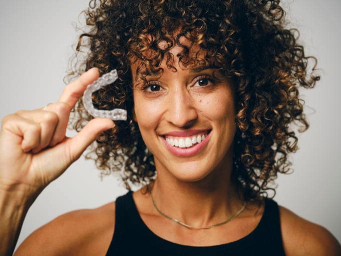 A woman smiling while holding her invisible braces retainer.