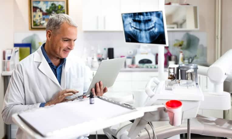A dentist is in his exam room with a patient's X-rays pulled up as he discusses treatment with them through teledentistry.