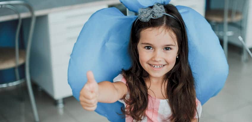 A young girl giving a thumbs up and smiling at the pediatric dentist's office.