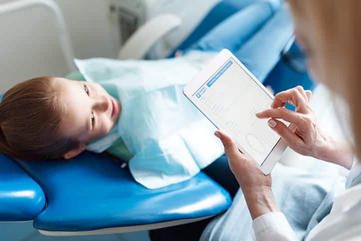 Focus is on a pediatric dentist filling out an electronic chart for their patient as she smiles while in the dental chair.