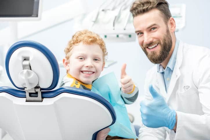 A young boy and a pediatric dentist both giving a thumbs up before a routine teeth cleaning.