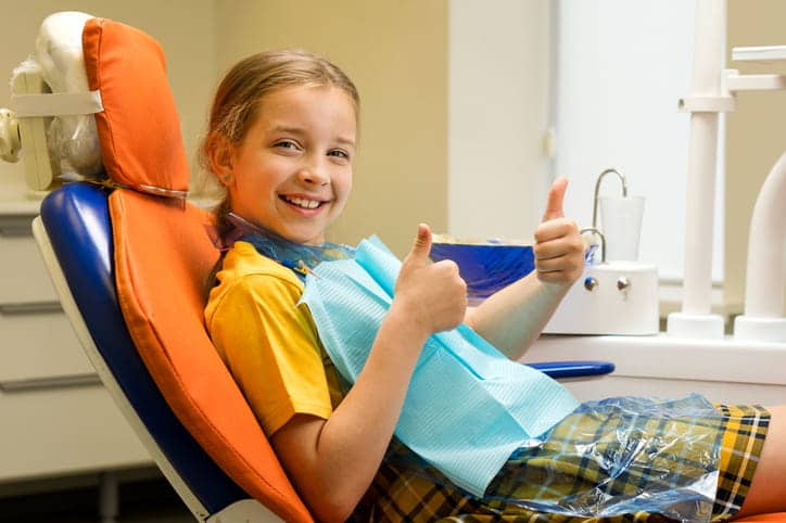 A young girl is giving two thumbs up while sitting in a dental chair after getting a dental sealant applied.