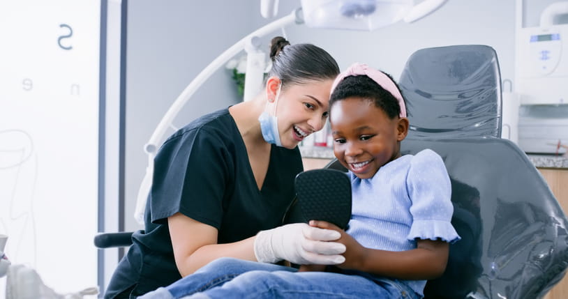 A dentist and a little girl smiling in a mirror together.