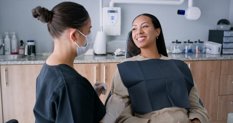 A dentist speaking to her client as she sits in the dental chair smiling.