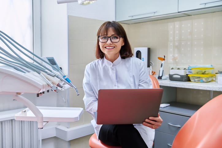 A dentist in a patient room smiling after speaking to them on her laptop during a teledentistry appointment. 