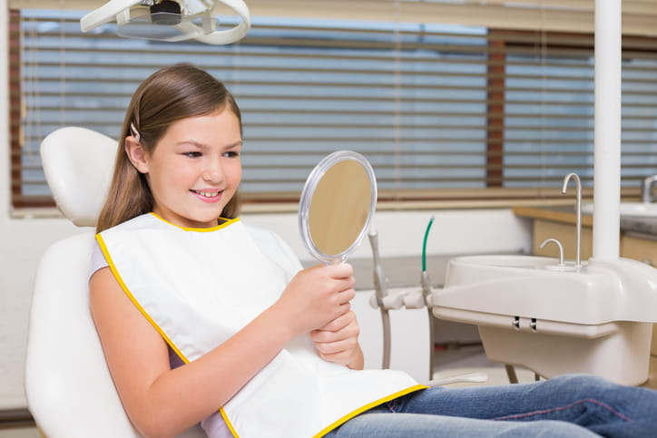 A young girl smiles as she looks into a handheld mirror at the dentist's office.