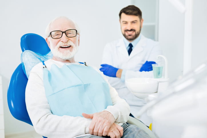 An elderly man smiling while sitting in a dental chair. His dentist is also smiling behind him. 