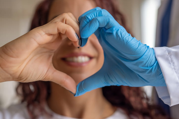 A dentist with one glove on forms a heart with her hands in front of her smile. 